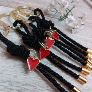 May include: Five black keychains with red heart charms. Each keychain is made of a black braided cord with a gold ring at the top. The cords are tied in a knot at the top and have a gold cap at the bottom.