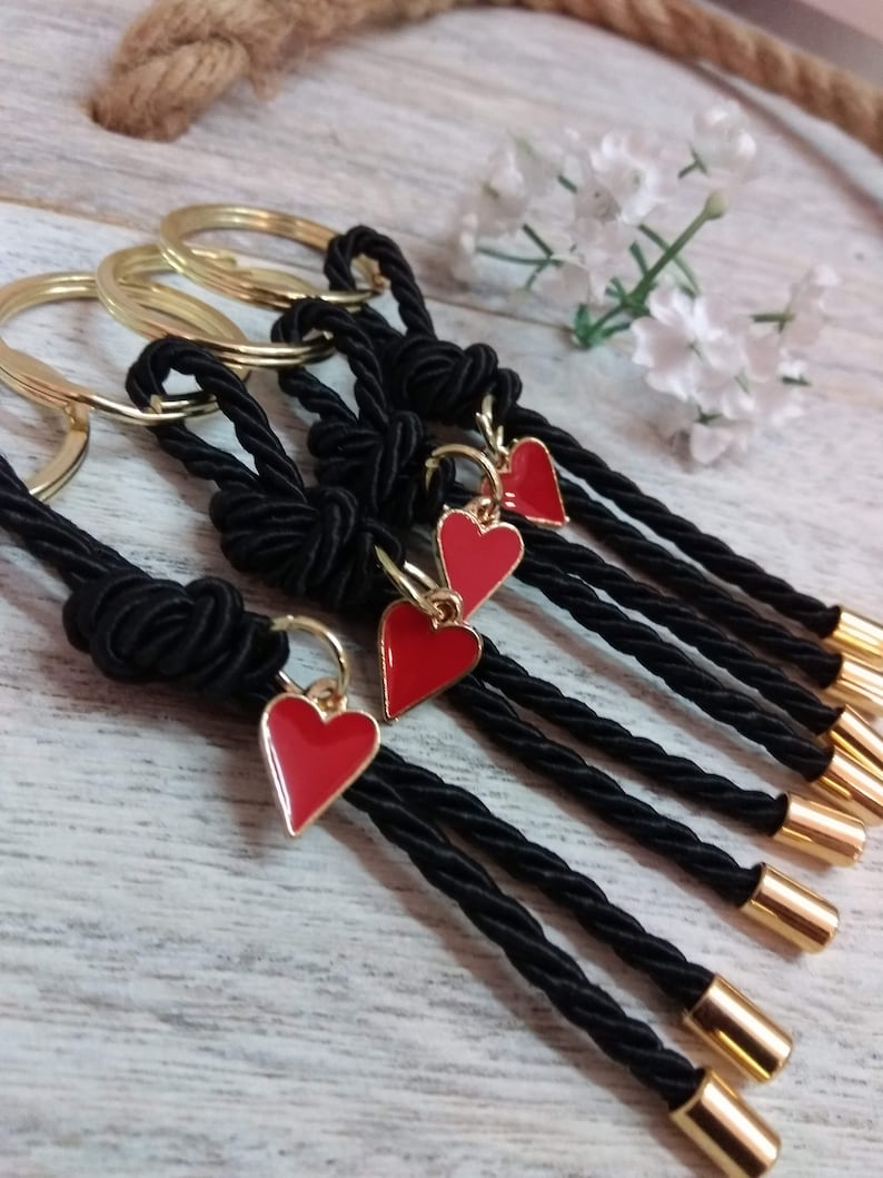 May include: Five black keychains with red heart charms. Each keychain is made of a black braided cord with a gold ring at the top. The cords are tied in a knot at the top and have a gold cap at the bottom.