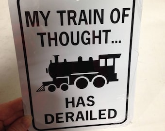 Può includere: Un cartello rettangolare bianco con bordo nero e la scritta "MY TRAIN OF THOUGHT... HAS DERAILED". Una silhouette nera di un treno è al centro. Il cartello ha un foro nella parte superiore per appenderlo.