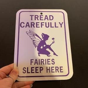Puede incluir: Un letrero de metal blanco con texto morado y una silueta morada de un hada. El letrero dice "Tread Carefully Fairies Sleep Here."