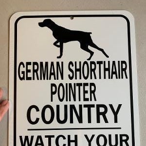 May include: White metal sign with black text and a silhouette of a German Shorthaired Pointer dog. The sign reads "German Shorthaired Pointer Country Watch Your Step!"
