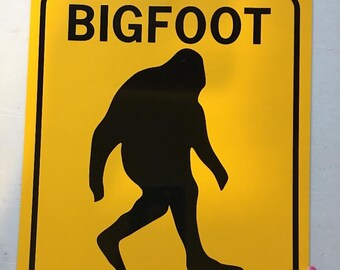 May include: Yellow warning sign with black text and a silhouette of Bigfoot walking. The sign reads "BIGFOOT XING".