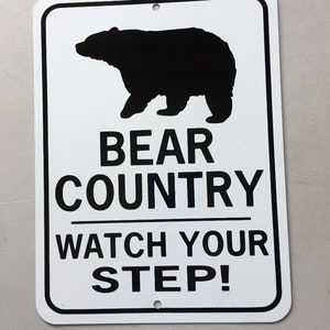 May include: White metal sign with a black silhouette of a bear. The sign reads "Bear Country Watch Your Step!"