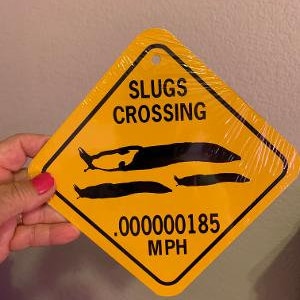 May include: Yellow diamond-shaped sign with black text that reads "Slugs Crossing" and ".000000185 MPH". The sign features a black silhouette of three slugs crawling across the sign.