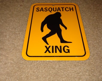 May include: Yellow warning sign with black text and a silhouette of a Sasquatch walking. The sign reads "Sasquatch Xing".