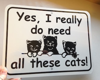 Puede incluir: Un letrero de metal blanco con texto negro e imágenes de tres gatitos negros. El letrero dice "Sí, realmente necesito todos estos gatos!"