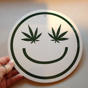 May include: White round sign with a green border and a green smiley face. The eyes of the smiley face are two green marijuana leaves.