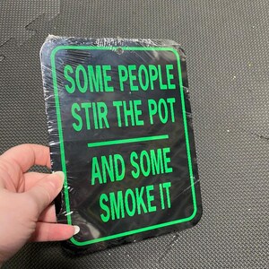 May include: Black and green metal sign with the text "Some people stir the pot and some smoke it".