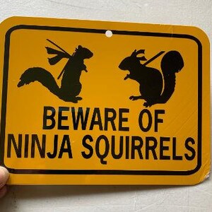 May include: Yellow warning sign with black text and silhouettes of two squirrels. The sign reads "Beware of Ninja Squirrels".
