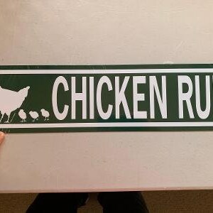 May include: A green metal sign with white text that reads "Chicken Run". A white silhouette of a hen and chicks is on the left side of the sign.