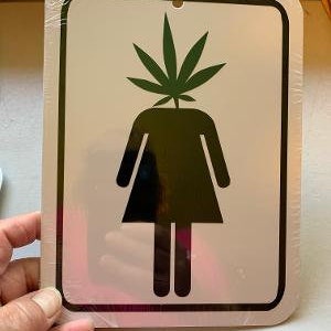May include: A white metal sign with a black border and a black silhouette of a person with a green marijuana leaf on their head. The sign is for a restroom.