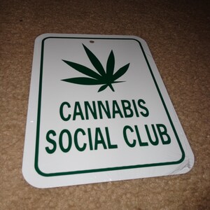 May include: White metal sign with green text and a green marijuana leaf. The sign reads "Cannabis Social Club".