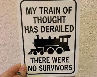 Può includere: Cartello in metallo bianco con testo nero e una silhouette nera di un treno. Il testo recita "Il mio treno di pensiero è deragliato. Non ci sono stati sopravvissuti."
