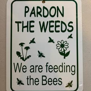May include: White sign with green text that reads "Pardon the Weeds We are feeding the Bees". The sign has a green silhouette of a dandelion, a daisy, and bees.