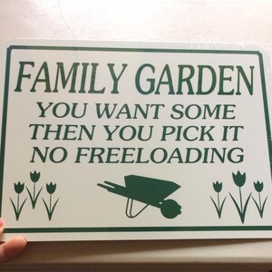 May include: White metal sign with green text that reads "Family Garden You Want Some Then You Pick It No Freeloading". The sign also features a green wheelbarrow and green tulips.