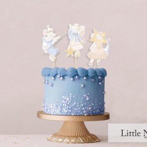 May include: A blue frosted cake with white sprinkles and three decorative toppers of mice wearing fairy wings and holding stars. The cake is on a gold cake stand.