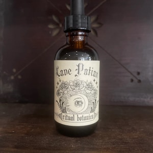 May include: A brown glass bottle with a dropper top and a white label that says "Love Potion" with a black and white illustration of a crescent moon, eye, and roses. The label also says "ritual botanica".