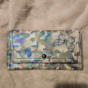 May include: A white wallet with a butterfly pattern and iridescent finish. The wallet has a snap closure and a silver button.
