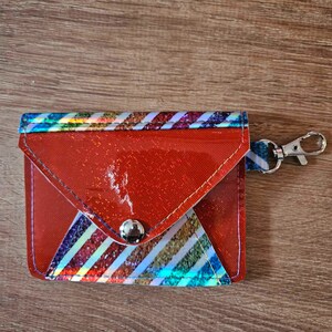 May include: A red envelope-style wallet with a rainbow striped pattern and a silver button closure. The wallet has a silver key ring attached.