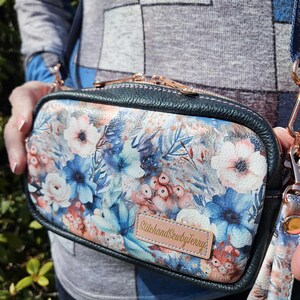 May include: A small, dark green crossbody bag with a floral print featuring blue, pink, and white flowers. The bag has a zipper closure and a gold-colored zipper pull. The bag has a small, brown leather tag with the text "Stitch and Sew by Terry" in gold lettering.