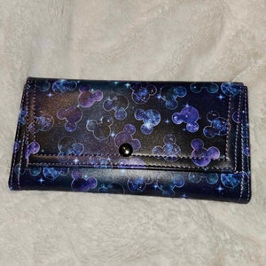 May include: A black wallet with a galaxy print featuring Mickey Mouse silhouettes. The wallet has a silver snap closure.