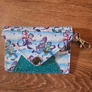 May include: A small blue and white fabric wallet with a teal glitter flap and a silver metal clasp. The wallet has a cartoon print of blue and purple figures with a white background. The wallet has a silver metal keychain attached.