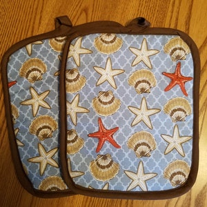 May include: A pair of blue and brown pot holders with a seashell and starfish pattern. The pot holders have a brown trim.