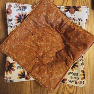 May include: Two square bowl cozies. The top cozy is a solid brown with a floral pattern. The bottom cozy is white with a turkey and floral design. Text on the bottom cozy reads "GOBBLE 'Til You WOBBLE!"