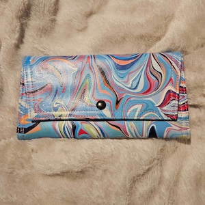 May include: A blue wallet with a colorful marbled pattern. The wallet has a snap closure and a black stitched seam.