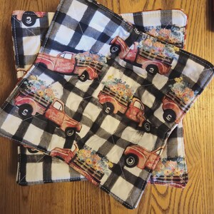 May include: Set of three quilted pot holders with a black and white buffalo check pattern. Each pot holder features a red vintage truck carrying a load of colorful flowers. The pot holders are square and have a stitched border.