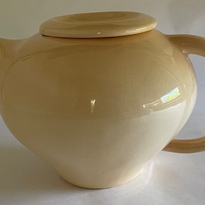 May include: Cream-colored ceramic teapot with a rounded body, curved handle, and a lid with a circular knob. The teapot has a glossy finish and a spout for pouring. A classic kitchenware item.