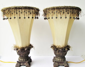 May include: A pair of ornate table lamps with beige fabric shades trimmed with brown fringe and black beads. The lamps have a brown, ornate base.