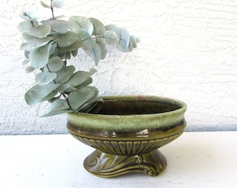 May include: A green glazed ceramic planter with a scalloped rim and a pedestal base. The planter is decorated with a swirl design and has a light green glaze on the inside. The planter is empty.