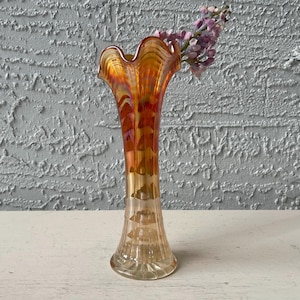 May include: An iridescent glass vase with a ruffled rim, displaying shades of orange, red, and gold. The vase has a slender, ribbed body and is filled with purple flowers. The vase is sitting on a white surface.