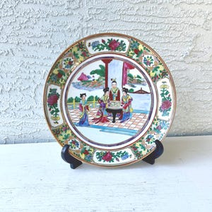 May include: A decorative porcelain plate with a detailed scene of figures in traditional attire. The plate features a gold rim and floral and foliate patterns. The central image depicts a gathering with a table, and the plate is displayed on a black stand.