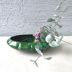 May include: A glossy, emerald green ceramic bowl with a textured exterior. The bowl holds a small arrangement of pink flowers and eucalyptus branches. The background is a textured white wall, creating a contrast with the bowl's color.