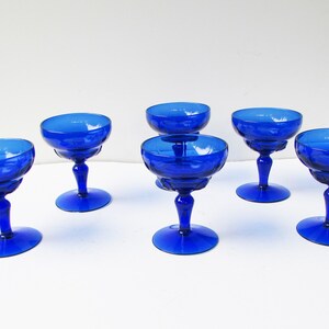 May include: Six cobalt blue glass champagne coupes with a fluted design. The coupes are arranged in a row on a white surface.
