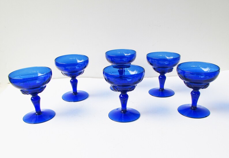 May include: Six cobalt blue glass champagne coupes with a fluted design. The coupes are arranged in a row on a white surface.