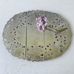 May include: A silver-colored, oval-shaped metal trivet with an intricate, openwork design. The trivet is divided into three hinged sections and features floral and scroll patterns. A sprig of purple flowers rests on the trivet.