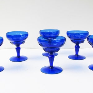 May include: Six cobalt blue glass champagne coupes with a fluted design. The coupes are arranged in a row on a white surface.