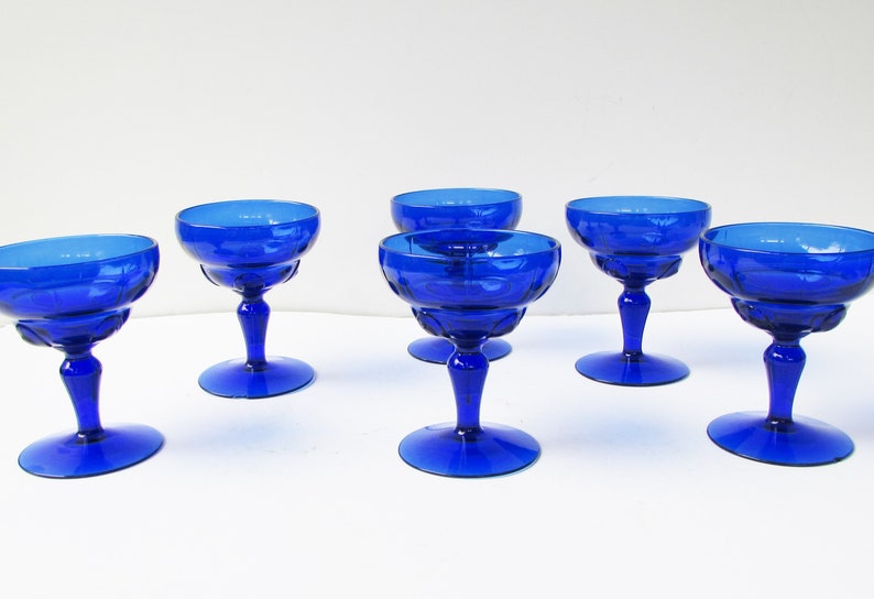 May include: Six cobalt blue glass champagne coupes with a fluted design. The coupes are arranged in a row on a white surface.