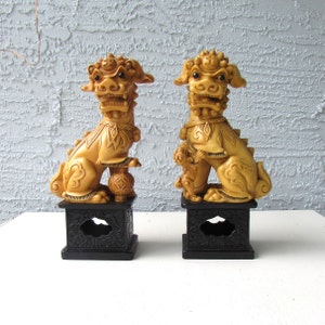 May include: A pair of decorative, carved, beige foo dogs, each standing on a black base with intricate carvings. The foo dogs are traditional Chinese guardian lions.