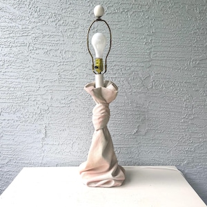 May include: A light pink table lamp with a unique knotted fabric design. The lamp features a white bulb, a brass-coloured lamp holder, and a white ceramic finial. The lamp is placed on a white surface against a textured grey wall.