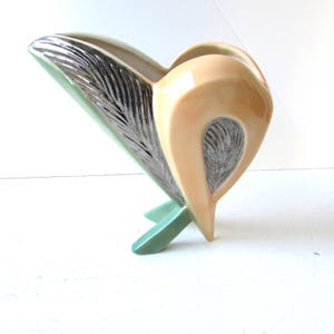 May include: A ceramic vase with a unique abstract design. The vase is a light beige colour with green and silver accents. It has a wide opening and a narrow base with three legs.