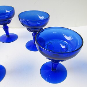 May include: Four cobalt blue glass stemmed dessert dishes with a decorative pattern. The dishes are on a white surface.