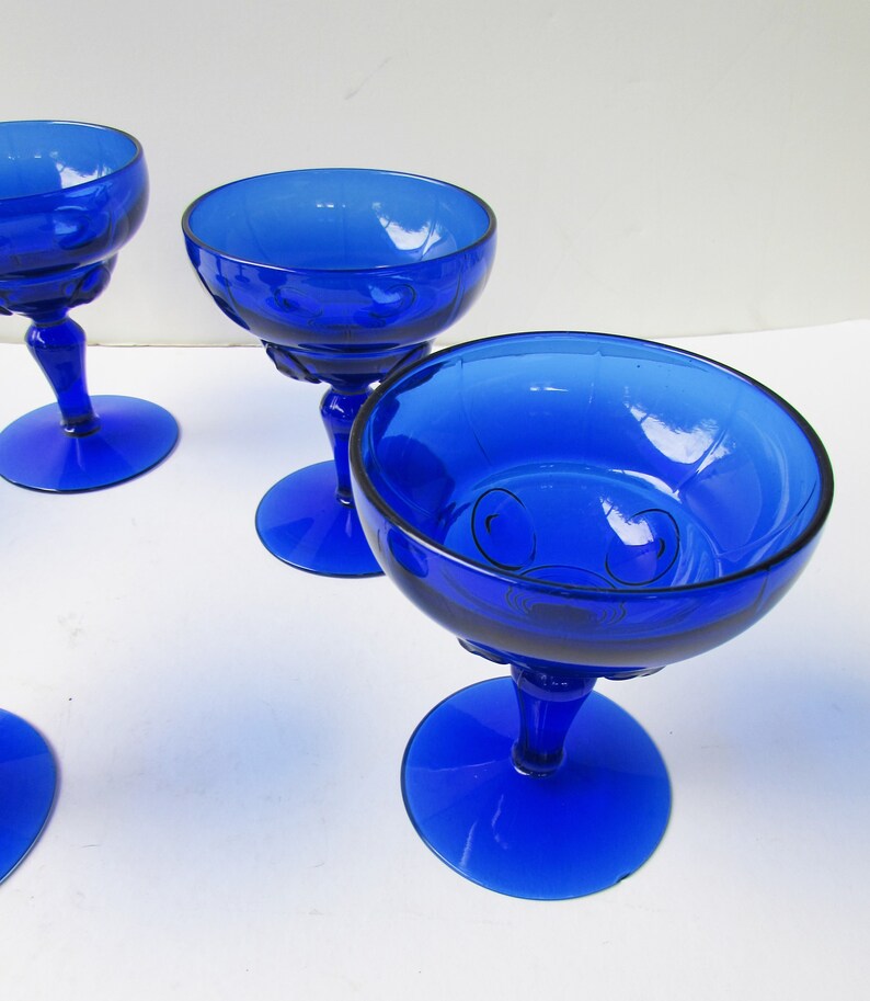 May include: Four cobalt blue glass stemmed dessert dishes with a decorative pattern. The dishes are on a white surface.