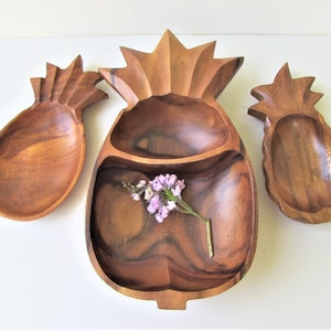 May include: Three wooden pineapple shaped serving bowls. The largest bowl has two sections and is decorated with a carved pineapple top. The other two bowls are smaller and have a single section. The bowls are made of dark brown wood.