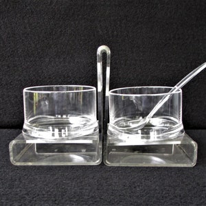 May include: A clear acrylic sugar and creamer set with a spoon. The set has two small bowls with a handle in the center. The bowls sit on a rectangular base.