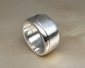 May include: A silver ring with a wide band and a textured design on the top. The ring has a brushed metal finish and a smooth, rounded edge. The ring is a piece of jewelry.