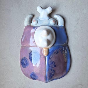 Könnte beinhalten: Eine Keramik-Wandskulptur in Form eines Käfers mit glänzender Oberfläche. Der Körper ist in Violett und Blau gehalten und mit dunkelblauen kreisförmigen Mustern versehen. Er hat einen weißen zentralen Vorsprung und einen weißen Kopf.